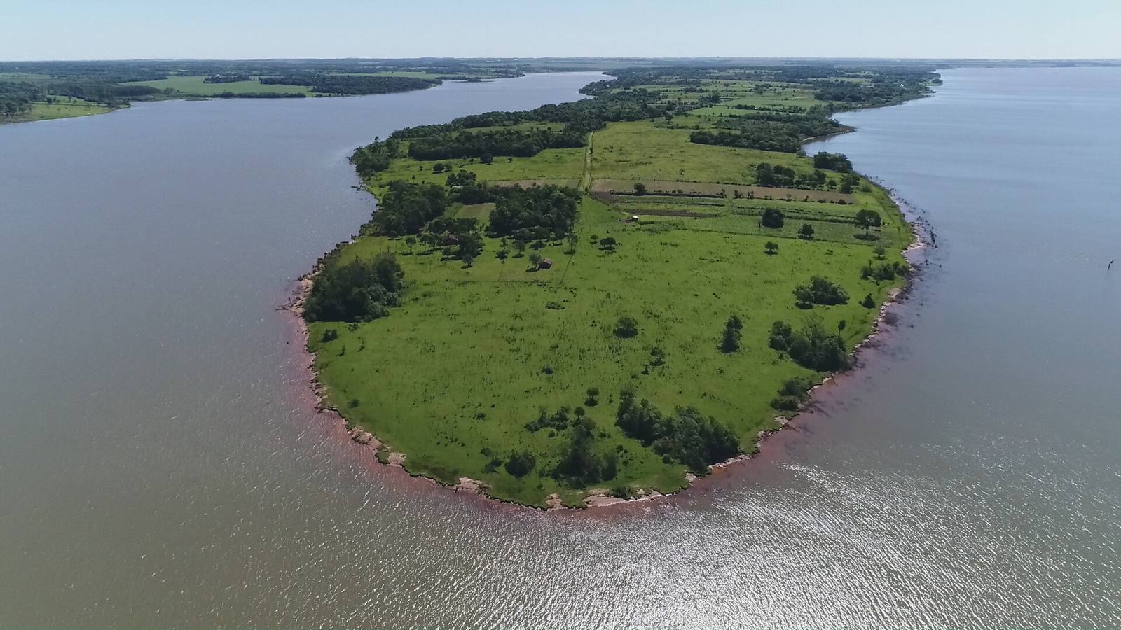Aerial view of the tip of the Zapallo peninsula: green fields, a red earth shore and the waters of Lake Yguazú on three sides.