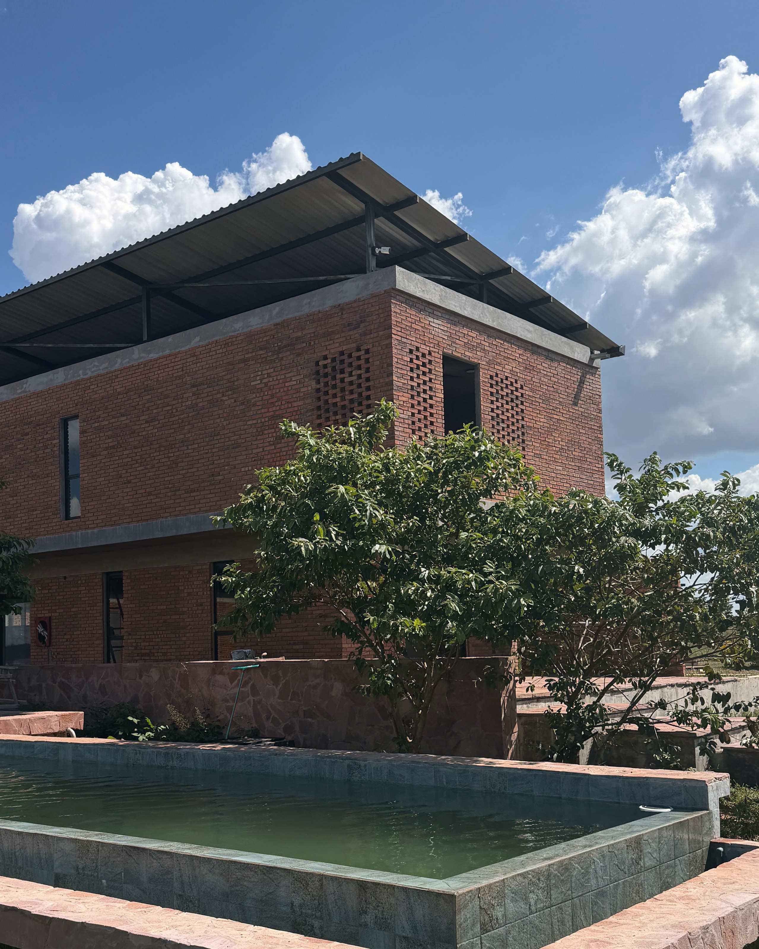 El edificio de ladrillo visto con muros de celosía, junto a la piscina de agua verde y borde de piedra rosada.